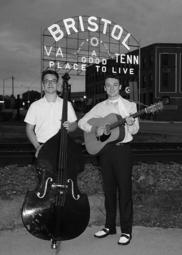 Zach McNabb holding a guitar and the Tennessee Esquires holding a cello in front of the Bristol, VA and TN sign