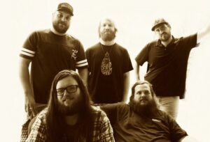 Three band members stand behind two other members that are sitting on the couch below