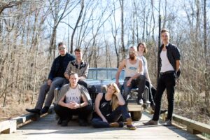 Members of the band (7 total) pose in front of old car on bridge