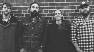 Four men standing against a brick wall. There is a black and white filter on the image.