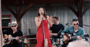 Woman in red dress singing into a microphone. Two men are sitting beside her, playing guitars. A man is behind her playing the drums.