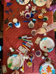 Arts and crafts spread across a table with a red tablecloth. 2 separate hands are seen reaching across the table to paint.