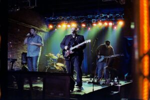 Three members in a band stand on stage with instruments and microphones in front of colorful lights