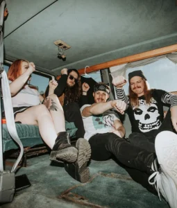 Five band members sit in a van dancing. Three men one women.