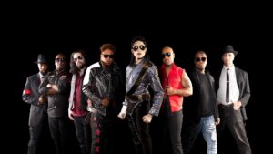 8 band members stand in front of a black background. The front center one is dressed like Michael Jackson