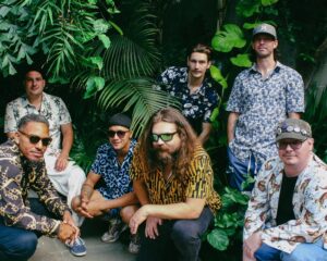 seven men dressed very exotically (unique out there kind of printed shirts) sitting in a jungle like place